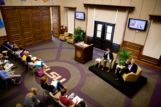Picture of a forum space with students in the audience and two men sitting in arm chairs in the front of room speaking to one another